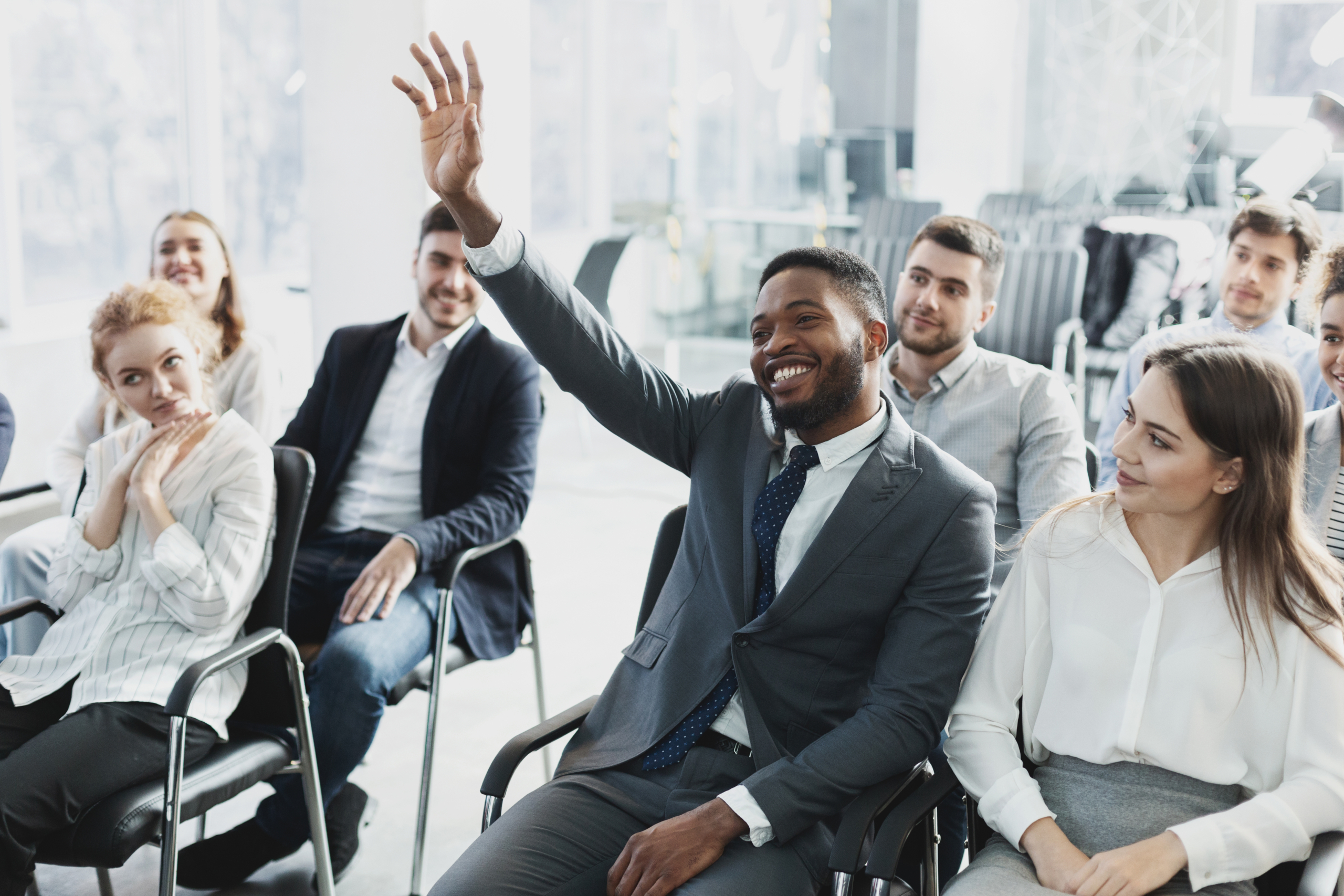 entire office having a meeting about their future relocation where one black man is raising hiss hand smiling to ask a question while his coworkers all look around smiling and inquisitive 