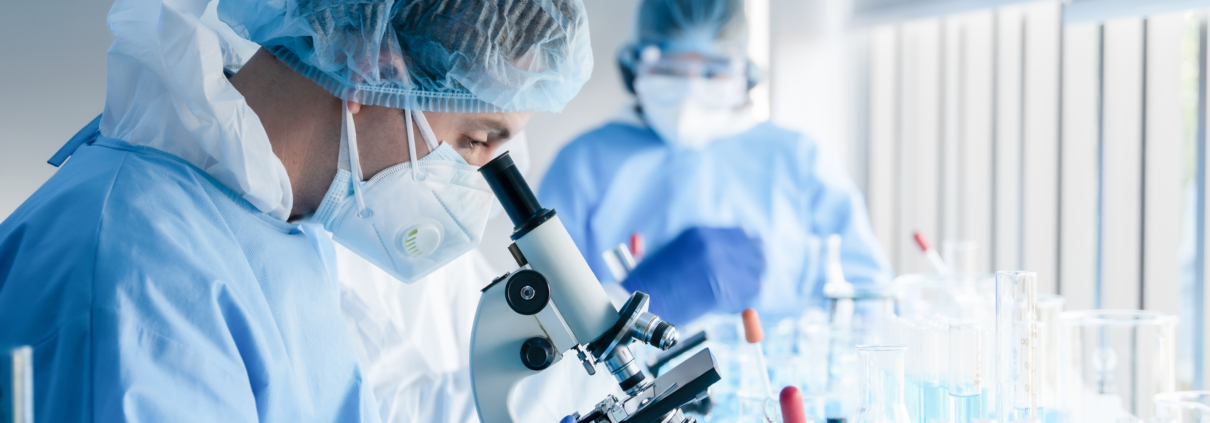 two scientists in a laboratory looking at a microscope wearing blue scrubs, surgical hair wraps and masks